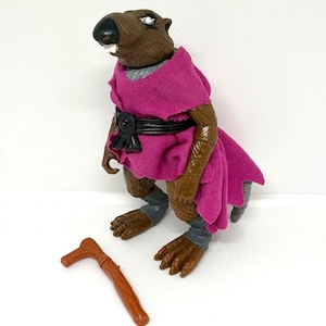 May include: A brown anthropomorphic rat action figure with white teeth and a black nose, wearing a magenta fabric robe with a black belt and grey leg wraps. A separate brown plastic walking stick accessory lies on the surface next to the figure.