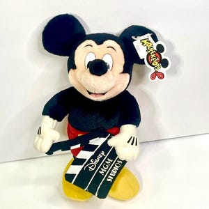 Puede incluir: Un peluche de Mickey Mouse, con pantalones cortos rojos, zapatos amarillos y guantes blancos, sostiene una claqueta de cine en blanco y negro. La claqueta dice "Disney MGM Studios". El juguete tiene una etiqueta "Mousekefun" adjunta a su oreja.