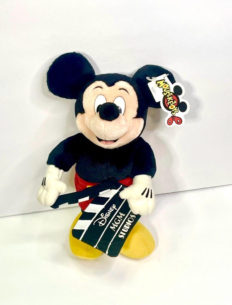 Puede incluir: Un peluche de Mickey Mouse, con pantalones cortos rojos, zapatos amarillos y guantes blancos, sostiene una claqueta de cine en blanco y negro. La claqueta dice "Disney MGM Studios". El juguete tiene una etiqueta "Mousekefun" adjunta a su oreja.