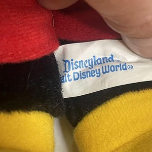 Puede incluir: Primer plano de un peluche con tela roja, negra y amarilla. Es visible una etiqueta con las palabras "Disneyland Walt Disney World". El dise&ntilde;o del juguete recuerda a un popular personaje de dibujos animados.