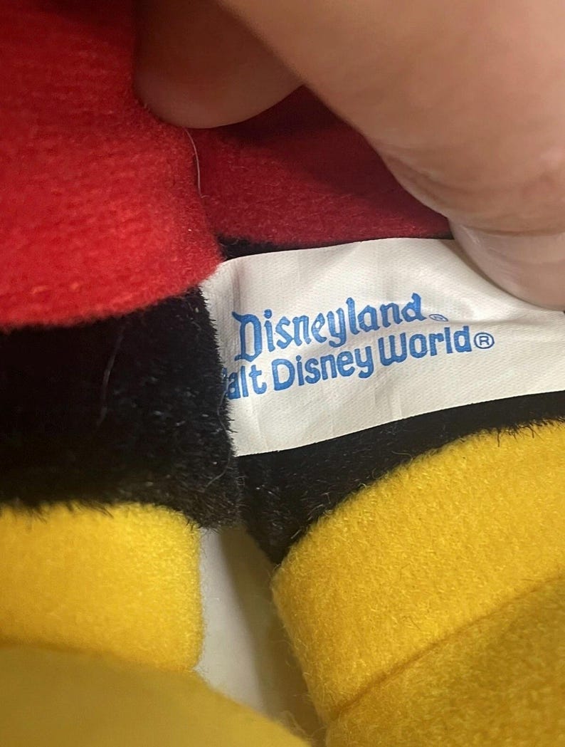 Puede incluir: Primer plano de un peluche con tela roja, negra y amarilla. Es visible una etiqueta con las palabras "Disneyland Walt Disney World". El dise&ntilde;o del juguete recuerda a un popular personaje de dibujos animados.
