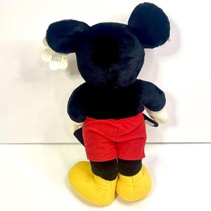 Puede incluir: Un peluche de Mickey Mouse, visto desde atr&aacute;s, con pelaje negro, pantalones cortos rojos, zapatos amarillos y guantes blancos. El juguete tiene grandes orejas negras y una etiqueta adjunta. Un personaje cl&aacute;sico de Disney.