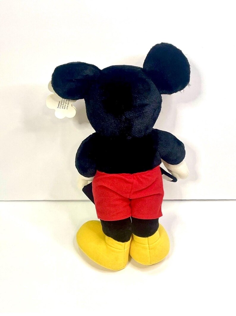 Puede incluir: Un peluche de Mickey Mouse, visto desde atr&aacute;s, con pelaje negro, pantalones cortos rojos, zapatos amarillos y guantes blancos. El juguete tiene grandes orejas negras y una etiqueta adjunta. Un personaje cl&aacute;sico de Disney.
