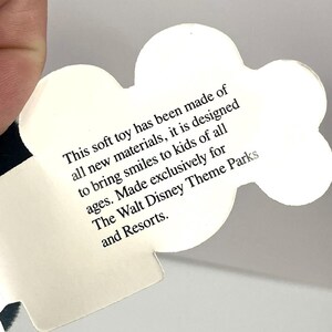 Puede incluir: Una etiqueta blanca en forma de nube con texto negro. El texto dice: "Este peluche ha sido fabricado con materiales nuevos, est&aacute; dise&ntilde;ado para sacar sonrisas a ni&ntilde;os de todas las edades. Fabricado exclusivamente para The Walt Disney Theme Parks and Resorts."