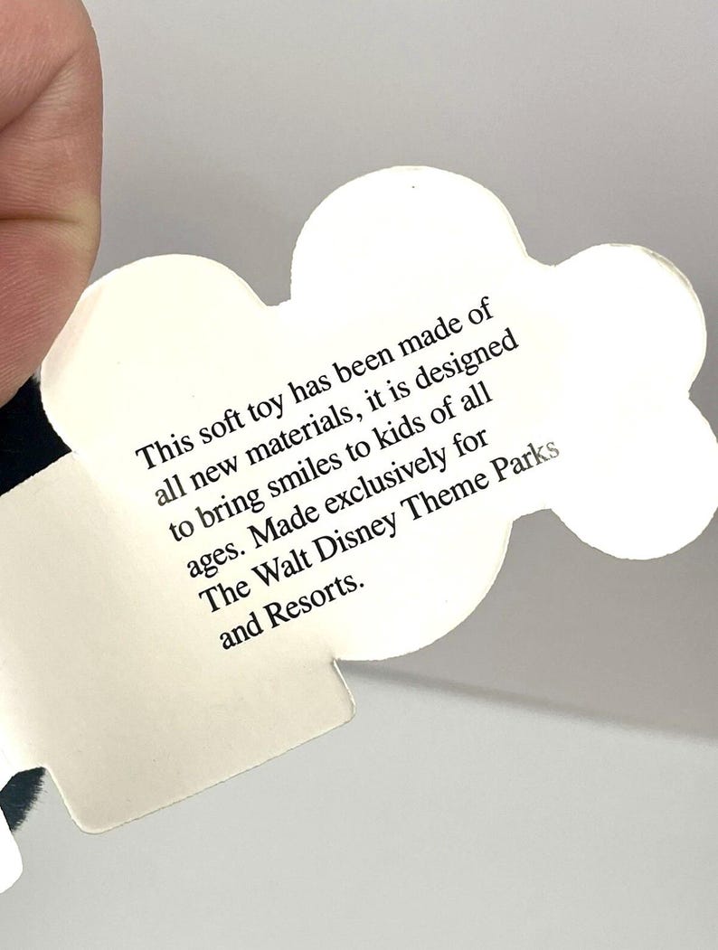 Puede incluir: Una etiqueta blanca en forma de nube con texto negro. El texto dice: "Este peluche ha sido fabricado con materiales nuevos, est&aacute; dise&ntilde;ado para sacar sonrisas a ni&ntilde;os de todas las edades. Fabricado exclusivamente para The Walt Disney Theme Parks and Resorts."