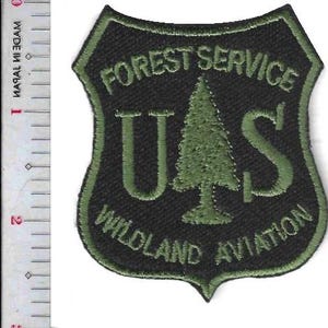 May include: A shield-shaped embroidered patch with a black background and olive green border. It features the text FOREST SERVICE, U.S with a pine tree icon, and WILDLAND AVIATION. A metal ruler shows the patch is approximately 3 inches tall.