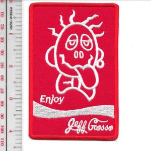 Pode incluir: Um patch retangular vermelho com um rosto de desenho animado bordado em branco, o texto "Enjoy" e "Jeff Grosso". O rosto do desenho animado tem os olhos fechados e a língua de fora. O patch tem aproximadamente 7,6 cm de altura.