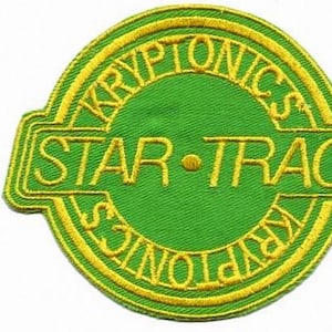 May include: Green and yellow embroidered patch with the words "KRYPTONICS" and "STAR-TRAC". The patch is circular with a rectangular border. The text is in yellow, and the background is green.