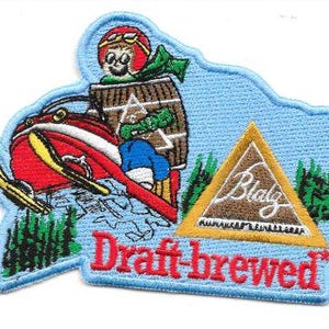 Op de afbeelding: Een geborduurde patch met een lichtblauwe achtergrond met een stripfiguur in een rode helm op een rode sneeuwscooter. Een bruin vat, een gouden driehoek met de tekst Blatz en de rode tekst Draft-brewed zijn zichtbaar. Een liniaal toont 9 cm.
