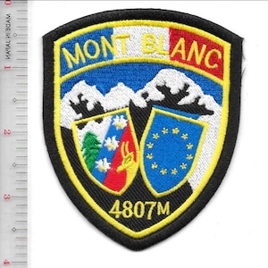 Könnte beinhalten: Schildförmiger Aufnäher mit den gelben Worten "MONT BLANC" über einer Bergkette. Der Aufnäher zeigt einen blauen Himmel, einen rot-weißen Abschnitt und zwei kleinere Embleme mit dem Text "4807M". Der Aufnäher hat einen schwarzen Rand und eine gelbe Umrandung.
