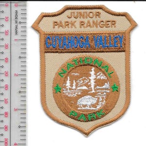 Puede incluir: Un parche bordado en forma de escudo con el texto JUNIOR PARK RANGER y CUYAHOGA VALLEY. Un emblema circular central representa un paisaje con árboles, montañas y un bisonte. Una regla muestra que el parche mide aproximadamente 9 cm.