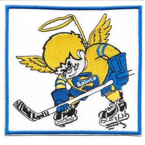 May include: Embroidered patch featuring a yellow angel playing hockey. The angel wears blue and yellow hockey gear and skates, holding a hockey stick. The word "Saints" is on the chest. The patch has a blue border and is on a white background.