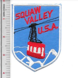 May include: Embroidered patch with a blue background featuring the text "SQUAW VALLEY U.S.A." in red. The patch depicts a cable car and mountain range in white, with a black cable car tower and red cable car.
