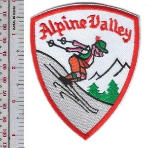 May include: Embroidered patch with a shield shape and a red border. The patch features the words "Alpine Valley" in red script above a skier in orange, pink, and green. Black mountains and green trees are also depicted.
