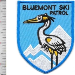 Peut inclure: Écusson brodé en forme de bouclier bleu. Le patch présente le texte "BLUEMONT SKI PATROL" au-dessus d'une illustration d'oiseau blanc et noir avec des accents jaunes, sur un fond bleu clair. Des sommets de montagnes blancs sont en bas.