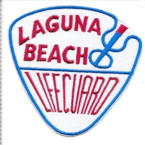 May include: A white embroidered patch with a blue border and the words "LAGUNA BEACH LIFEGUARD" in red. A blue and red rescue buoy graphic is on the right side of the patch.