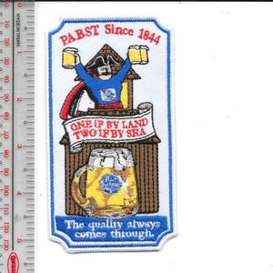 Może przedstawiać: Haftowana naszywka z logo Pabst Blue Ribbon. Projekt zawiera postać z kreskówki trzymającą kufle piwa, napis "PABST Since 1844" oraz frazę "ONE IF BY LAND TWO IF BY SEA". Naszywka ma niebieską obwódkę.