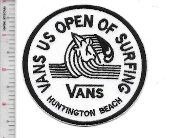 Könnte beinhalten: Runder Aufnäher mit schwarzem Rand und weißem Hintergrund. Der Text "VANS US OPEN OF SURFING" ist kreisförmig angeordnet. In der Mitte befindet sich ein stilisiertes Wellen- und Surfer-Design mit dem Wort "VANS" darunter und "HUNTINGTON BEACH" unten.