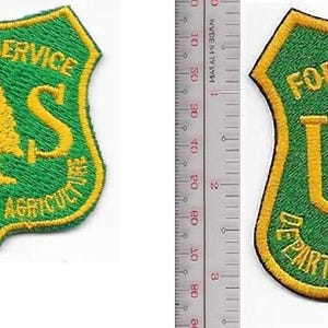 Puede incluir: Un parche bordado en forma de escudo con fondo verde y borde amarillo. Presenta el texto FOREST SERVICE y DEPARTMENT OF AGRICULTURE en amarillo, con un pino amarillo central y las letras U y S. Una regla muestra una altura de 5 cm.