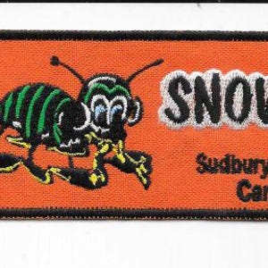May include: An orange rectangular patch with a black border features a cartoon bug with green and black stripes, and the word "SNOWBUG" in white. Below the text is "Sudbury, Ontario Canada".