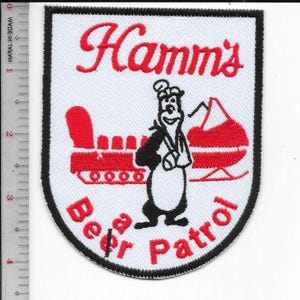 Könnte beinhalten: Weißer Aufnäher mit schwarzem Rand mit dem Text "Hamm's" in roter Schrift. Ein Cartoon-Bär in schwarzer Kleidung befindet sich in der Mitte, mit einem roten Zug und einer roten Struktur auf beiden Seiten. Die Worte "Beer Patrol" sind rot.