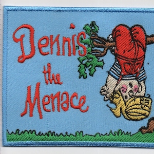 May include: A rectangular embroidered patch with a light blue background featuring the red text Dennis the Menace. A cartoon character in red overalls hangs upside down from a brown tree branch. A ruler shows the patch is 3.5 inches wide.