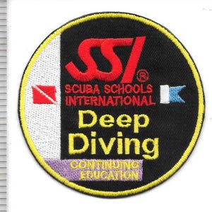 Könnte beinhalten: Ein kreisförmiger gestickter Aufnäher mit schwarzem Hintergrund und gelbem Rand. Er zeigt das rote SSI-Logo sowie den Text Scuba Schools International, Deep Diving und Continuing Education. Ein Lineal zeigt, dass der Aufnäher ca. 9 cm breit ist.
