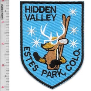 May include: Embroidered patch with a blue shield design featuring the text "HIDDEN VALLEY ESTES PARK, COLO." A cartoon deer wearing a hat and holding skis is in the center, surrounded by white starbursts. The patch has a black border.