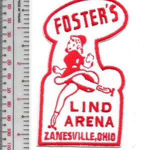 Könnte beinhalten: Ein weißer gestickter Aufnäher mit rotem Rand und rotem Text FOSTER'S LIND ARENA ZANESVILLE, OHIO, mit einer zentralen Grafik einer Figur auf Schlittschuhen. Ein Lineal links zeigt eine Höhe von 10 cm an.