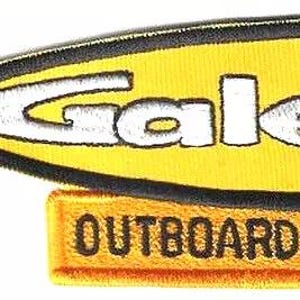 Può includere: Una toppa ricamata a forma di gagliardetto giallo con bordo nero e la scritta bianca Gale, posizionata sopra una barra rettangolare gialla con la scritta nera OUTBOARD MOTORS. Un righello mostra che la toppa è larga circa 5,7 cm.