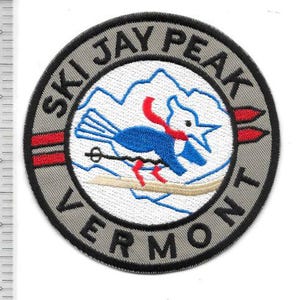 Può includere: Toppa ricamata circolare con la scritta "SKI JAY PEAK VERMONT". Il design presenta un uccello blu con una sciarpa rossa che scia su un pendio innevato, con una catena montuosa sullo sfondo.