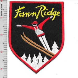 May include: Embroidered patch with a black background and a red border. The patch features the text "Fawn Ridge" in yellow, a skier in red and white, and brown skis. White pine trees are at the bottom.