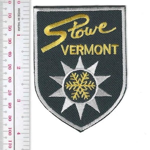 Könnte beinhalten: Ein schildförmiger Aufnäher mit den Worten "Stowe Vermont" in goldener Schrift. Unter dem Text befindet sich ein silbernes und goldenes Schneeflocken-Design. Der Aufnäher hat einen schwarzen Hintergrund mit silberner Umrandung.