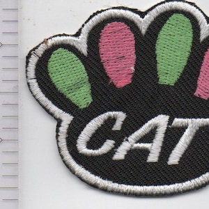 May include: A black embroidered patch shaped like a paw print with alternating light green and pink toe pads. The word CAT is stitched in white thread across the center. A ruler is visible on the left, showing the patch is approximately 2 inches wide.