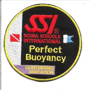 Puede incluir: Un parche bordado circular con fondo negro y borde amarillo. Presenta el logotipo rojo de SSI, el texto Scuba Schools International, Perfect Buoyancy y Continuing Education. Una regla muestra que el parche mide aproximadamente 9 cm de alto.