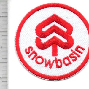 Może przedstawiać: Okrągła naszywka z białym środkiem i czerwoną obwódką. Środek zawiera czerwony, stylizowany wzór strzałki nad słowem "snowbasin" w kolorze czerwonym, wszystko na białym tle. Do odzieży.