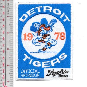 May include: A rectangular patch with a blue border and white background. The patch features the words "DETROIT TIGERS" in white, surrounding a cartoon tiger holding a baseball bat. The numbers "1978" are in orange. The patch also includes the text "OFFICIAL SPONSOR" and "Stroh's BEER".