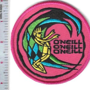 Peut inclure: Un écusson rond brodé rose avec un motif de surfeur. Le design comprend une figurine jaune, des accents bleus et verts, et le texte "O'NEILL" en noir. L'écusson a une bordure rose.