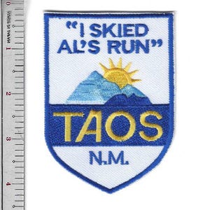 May include: A shield-shaped patch with a white background and blue border. The patch features the text "I SKIED AL'S RUN" above a mountain and sun graphic. Below is "TAOS N.M." in yellow lettering.