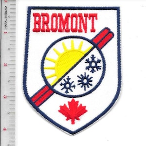May include: Embroidered patch with the word "BROMONT" in red. The patch features a sun and snowflakes, bisected by a red stripe. A red maple leaf is at the bottom, all within a white shield with a blue border.