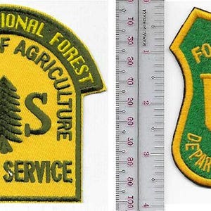May include: Two embroidered patches next to a metal ruler. The left patch is yellow with green text reading ANGELES NATIONAL FOREST DEPARTMENT OF AGRICULTURE FOREST SERVICE and a tree. The right patch is green with yellow text reading FOREST SERVICE U S DEPARTMENT OF AGRICULTURE and a tree.