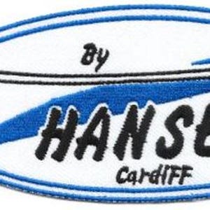 Puede incluir: Parche bordado con fondo ovalado blanco y borde azul. El parche presenta un diseño de tabla de surf negra y las palabras "HANSEN CARDIFF" en negro, con "by" encima de la tabla de surf. Un diseño de ola azul está detrás de la palabra "HANSEN".