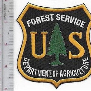 Puede incluir: Un parche bordado en forma de escudo con el texto FOREST SERVICE y DEPARTMENT OF AGRICULTURE en blanco, y un árbol verde entre las letras U y S en dorado. Una regla metálica muestra que el parche mide aproximadamente 9 cm de altura.