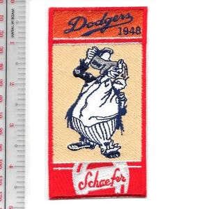 May include: A rectangular patch with a cartoon figure in a white outfit with blue accents, holding a beer stein. The patch has a red border and includes the text "Dodgers 1948" and "Schaefer" in cursive.