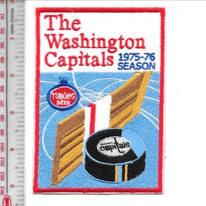 Può includere: Toppa ricamata con "The Washington Capitals" in rosso e "1975-76 SEASON". Presenta un disco da hockey, un logo della birra e un design di stecca da hockey. La toppa ha un bordo rosso e uno sfondo blu.