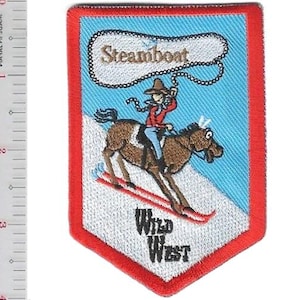 Puede incluir: Parche bordado con temática del Salvaje Oeste. El parche presenta a un vaquero montando a caballo sobre esquís, con la palabra "Steamboat" arriba y "Wild West" abajo. El parche tiene un borde rojo y un fondo azul.