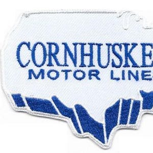 Op de afbeelding: Een witte geborduurde patch in de vorm van een kaart van de Verenigde Staten met blauwe tekst CORNHUSKER MOTOR LINES. De patch ligt naast een liniaal die een breedte van ongeveer 9 cm en een hoogte van 6,5 cm aangeeft.
