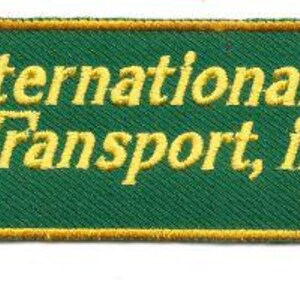 May include: A rectangular green patch with rounded corners. The patch features the words "International Transport, inc." in yellow, with a yellow border. The text is embroidered on the patch.