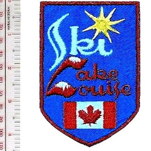 Könnte beinhalten: Ein schildförmiger, bestickter Aufnäher mit blauem Hintergrund, der den Text 'Ski Lake Louise' in Weiß und Rot, eine gelbe Sonne und die kanadische Flagge unten zeigt. Ein Lineal zeigt, dass der Aufnäher etwa 9 cm hoch ist.