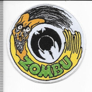 Puede incluir: Un parche bordado circular que presenta una criatura amarilla con cara de calavera y cabello negro, una silueta de murciélago negra central y el texto verde ZOMBU. Una regla a la izquierda muestra un diámetro de aproximadamente 9 cm.